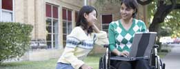 Girl sitting with a handicap girl that is in a wheelchair with a laptop in her lap